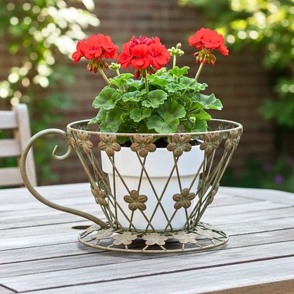 Rustic metal teacup shaped plant stand with flower wirework detail holding a red geranium on a garden table